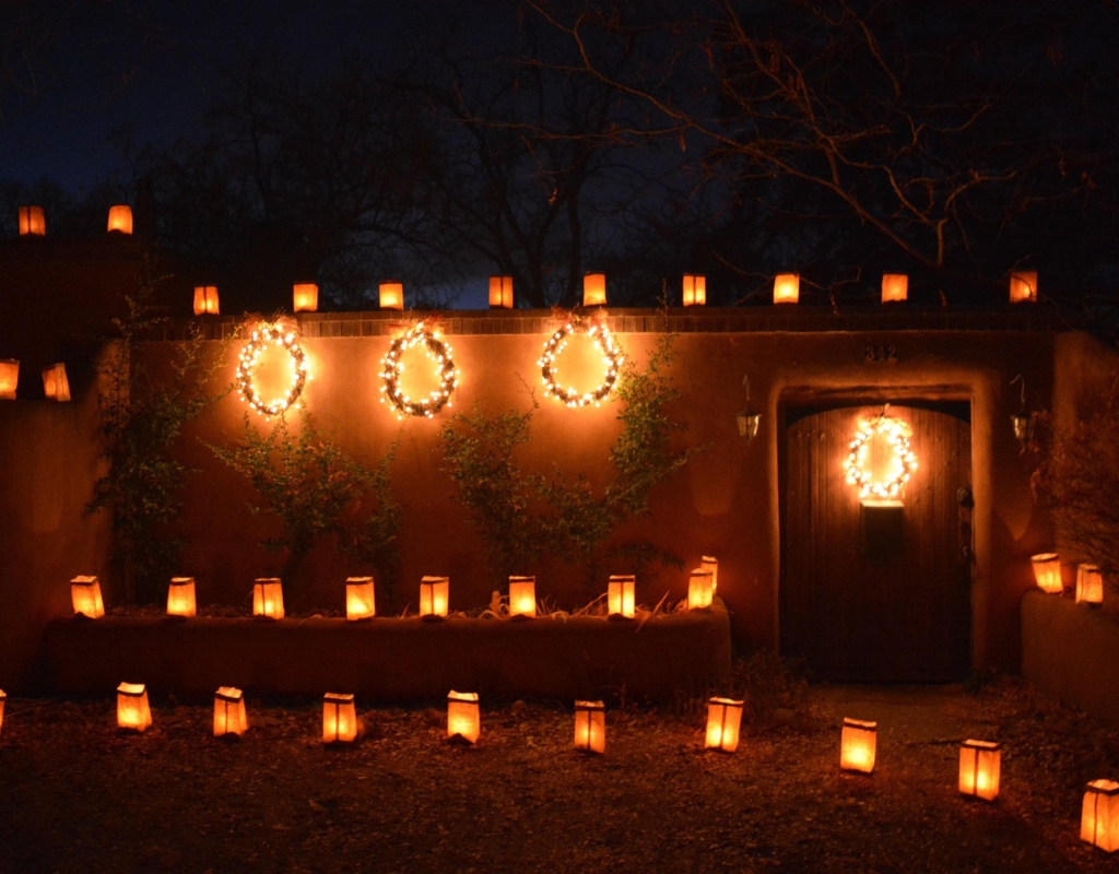 A wall with a wooden doorway where farolitos light up the darkness. Along the wall are also some wreaths wrapped in bright lights.