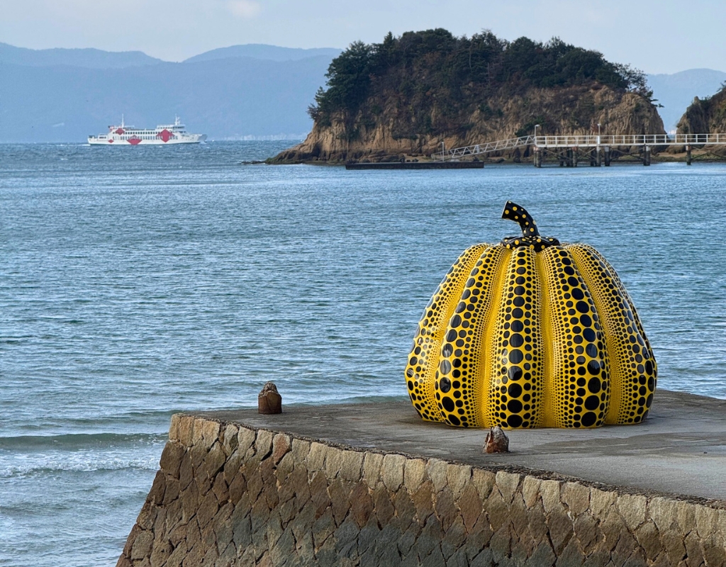 Photograph of a sculpture of a yellow pumpkin with black dots sitting on a concrete pier near the ocean. Behind the pumpkin, water extends with a small island on the horizon and a boat can be seen in the background.