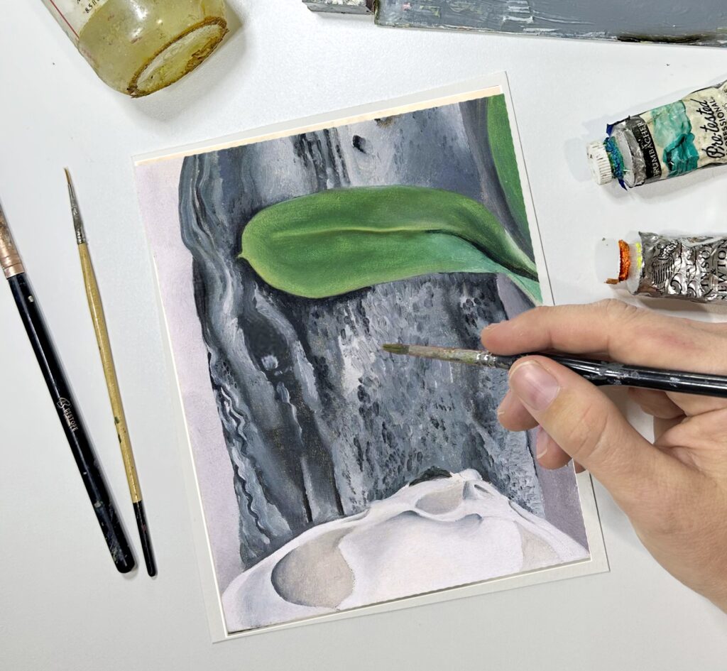 Photograph from above of a painting of a shell against a grey background with a green leaf above. Hovering over the painting is a hand with a small paintbrush. Nearby are paint tubes and brushes.