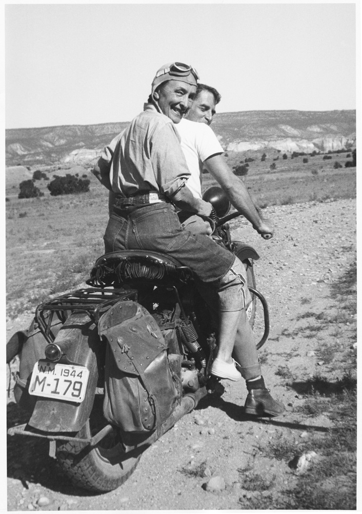 Georgia O'Keeffe on the back of a motorcycle holding on to a male friend (Maurice Grosser) who is driving. She is turning back looking towards the camera, in an expansive New Mexican landscape. She is wearing denim jeans, a chambray shirt, a cap with goggles, and sneakers.