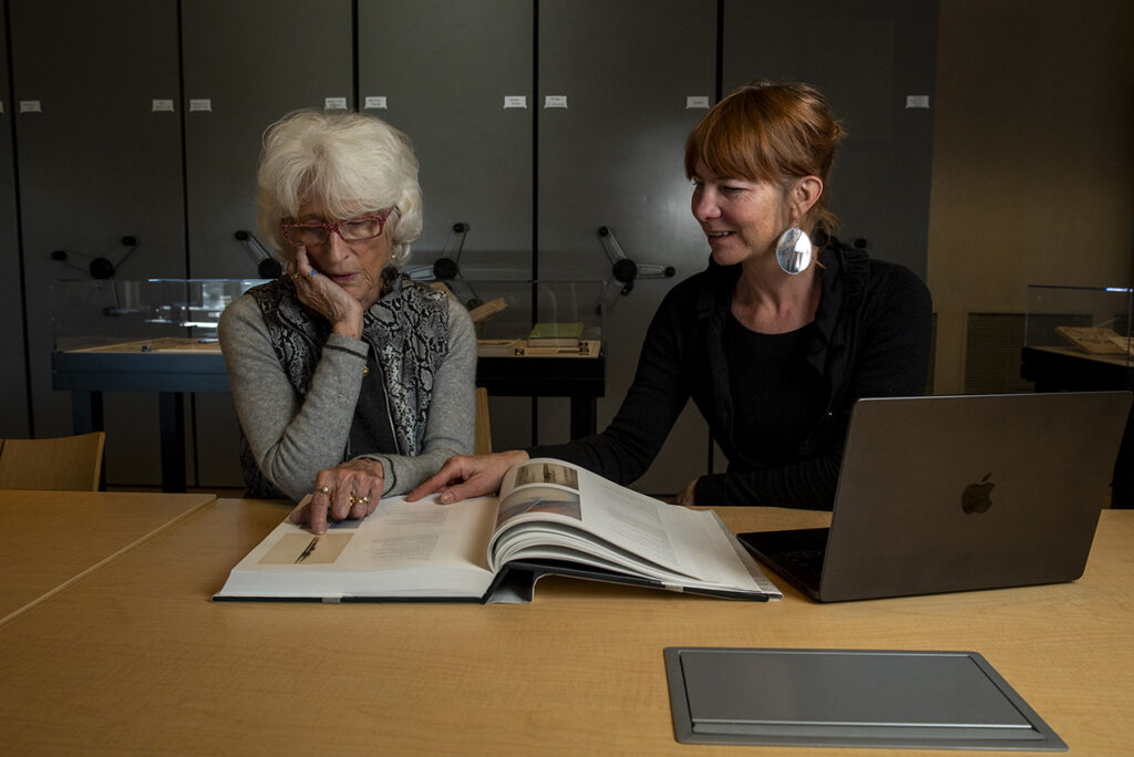 Photograph of two people sitting at a table looking at a large book. 