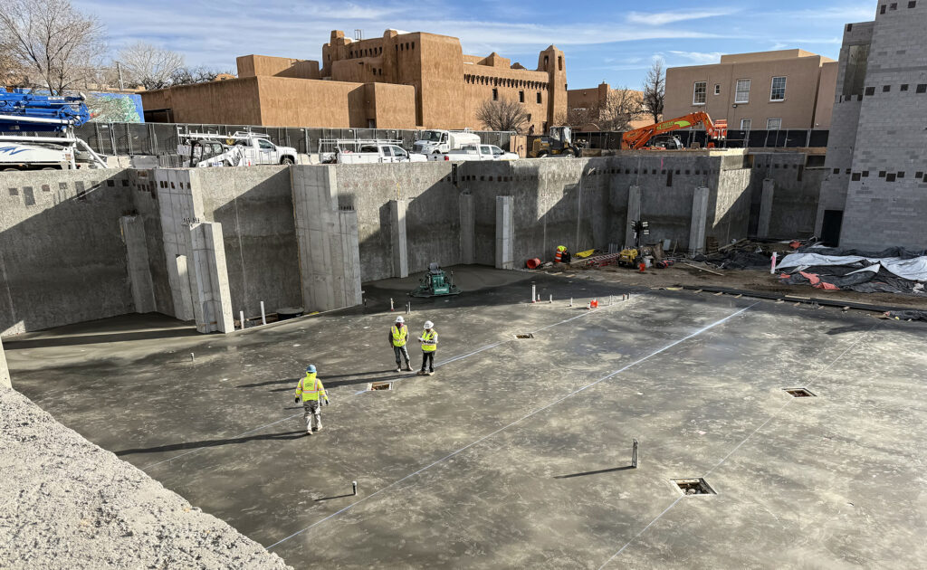 Photograph of a large construction site, where a concrete foundation and walls have been built. At the center of the site a few workpeople with hard hats and bright vests stand. Above the site an adobe building is visible in the background.