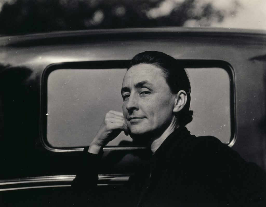 Quarter length portrait of O'Keeffe dressed in black with her hair pulled back leaning against a car. She looks at the camera with her face in semi-profile wearing a slight smile with her hand leaning against her cheek.