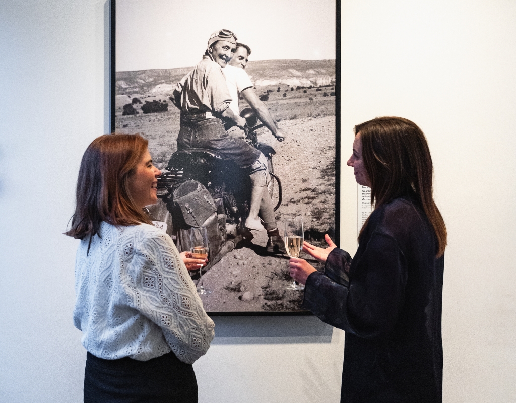 Two people holding glasses standing in front of a large photograph of Georgia O'Keeffe riding the back of a motorcycle.