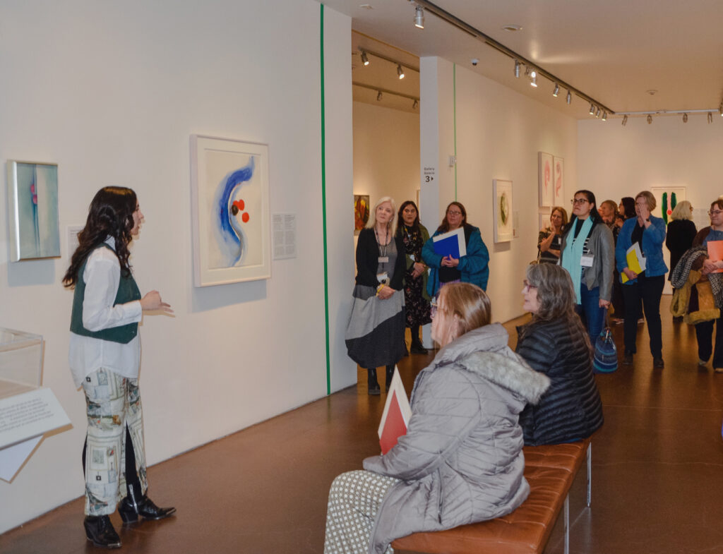 A group of people stand and sit in a gallery while a person speaks to the group.