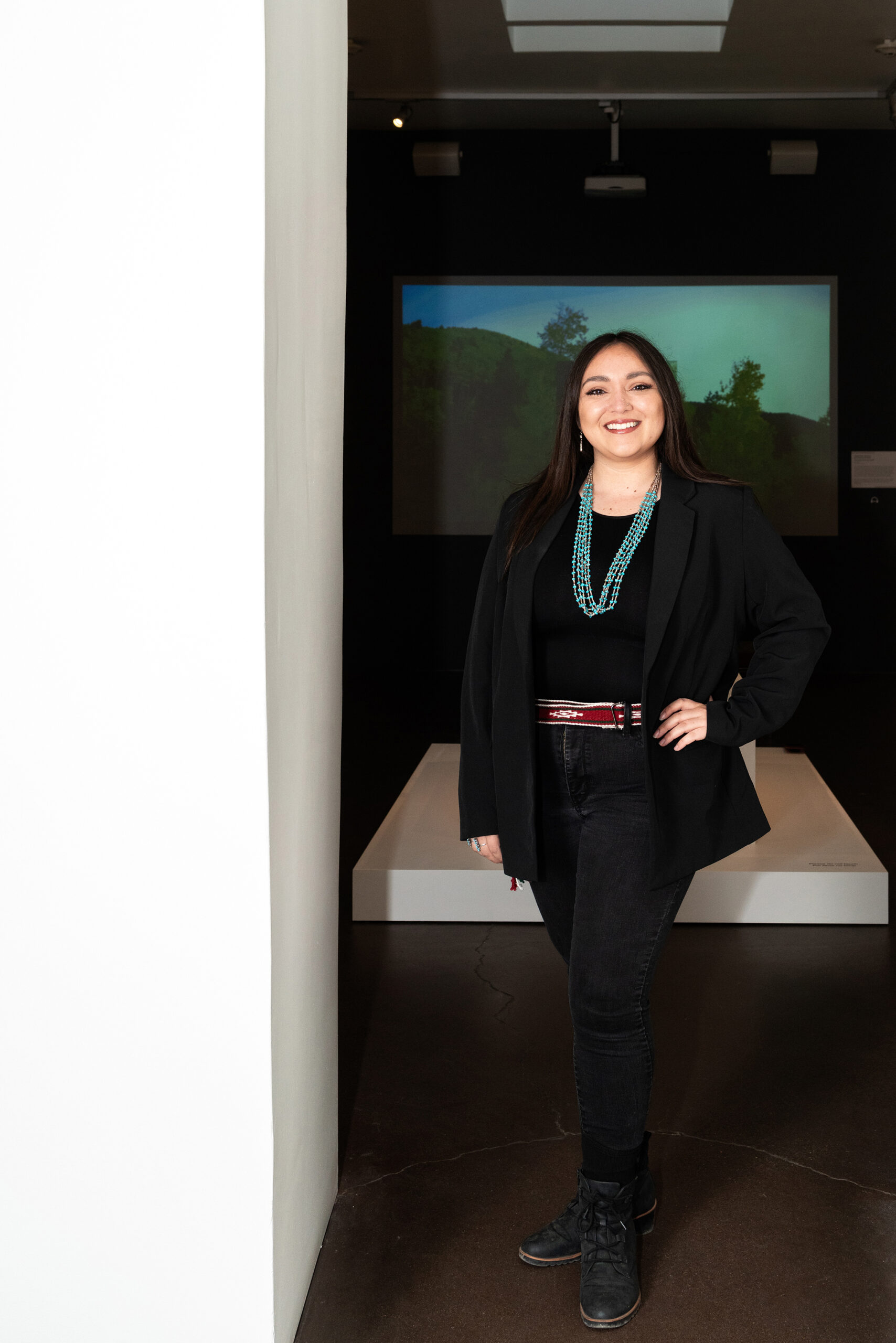Photograph of a person with long dark hair smiling and standing in front of a screen with a movie playing in the background.
