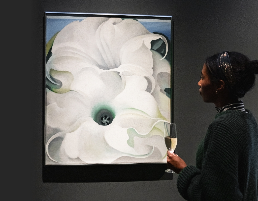 Photograph of a person with their hair tied up standing in front of a large painting of white flowers against a dark wall. In their hand they hold a filled glass.