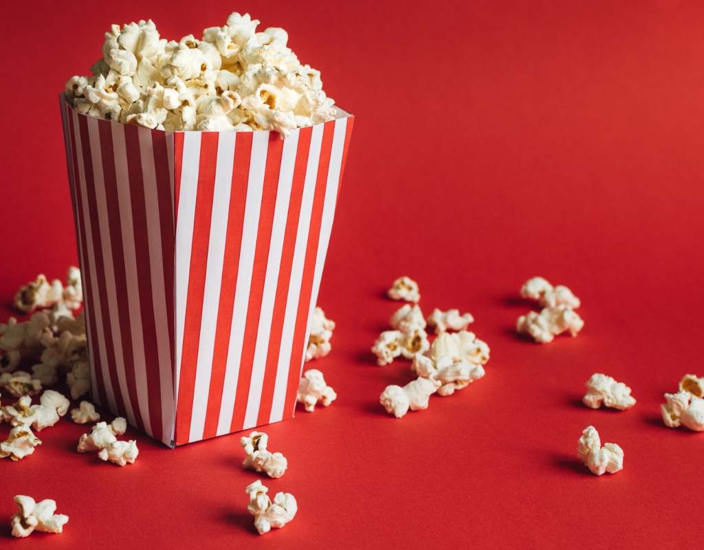 A pin-stripped container holding popcorn against a red background. Some popcorn pieces are also strewn about.