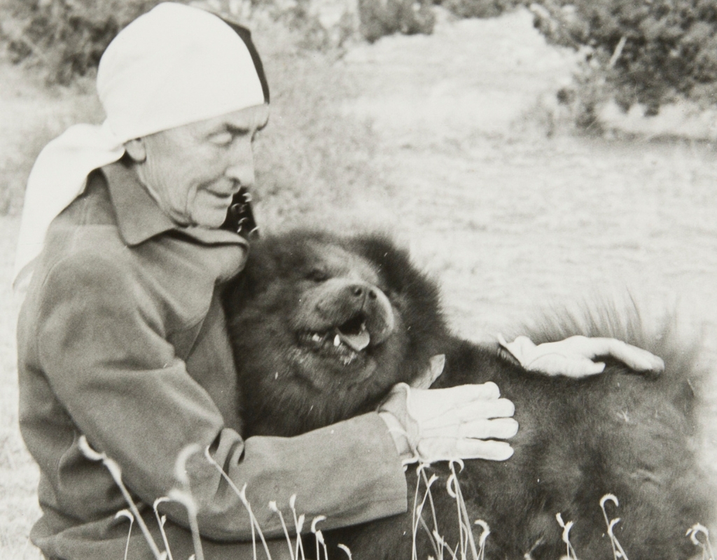 Black and white photograph of Georgia O'Keeffe wearing a jacket and a scarf wrapped around her head. O'Keeffe sits on the ground embracing a Chow Chow dog.