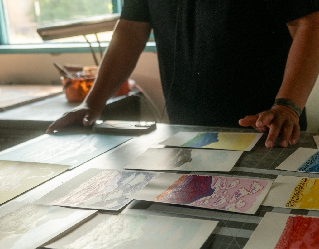 A photograph of a person's hands hovering over several small paintings of a flat-topped mountain laid on a table.