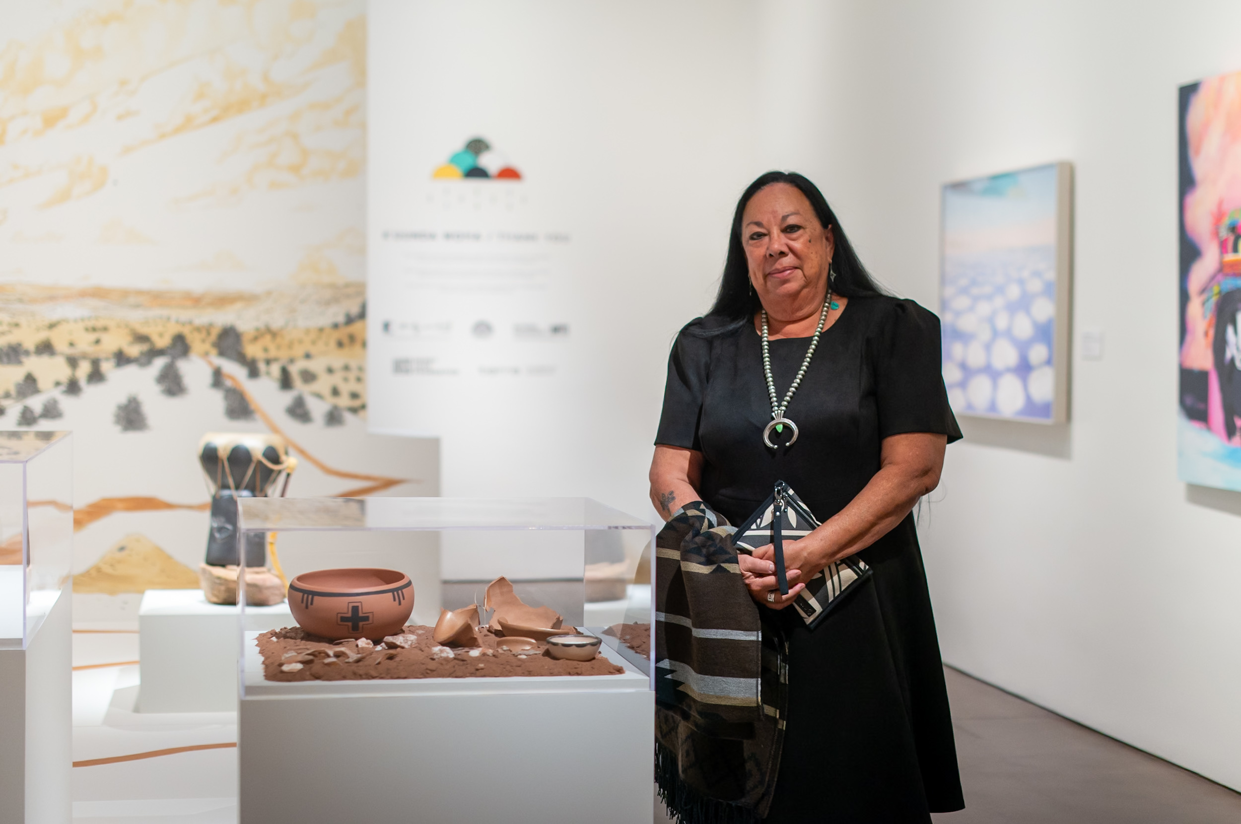 Photograph of a person with long dark hair in a long dark dress wearing a silver necklace and standing in front of a display case containing a clay pot surrounded by earth and other pottery shards. They stand in a gallery with white walls.
