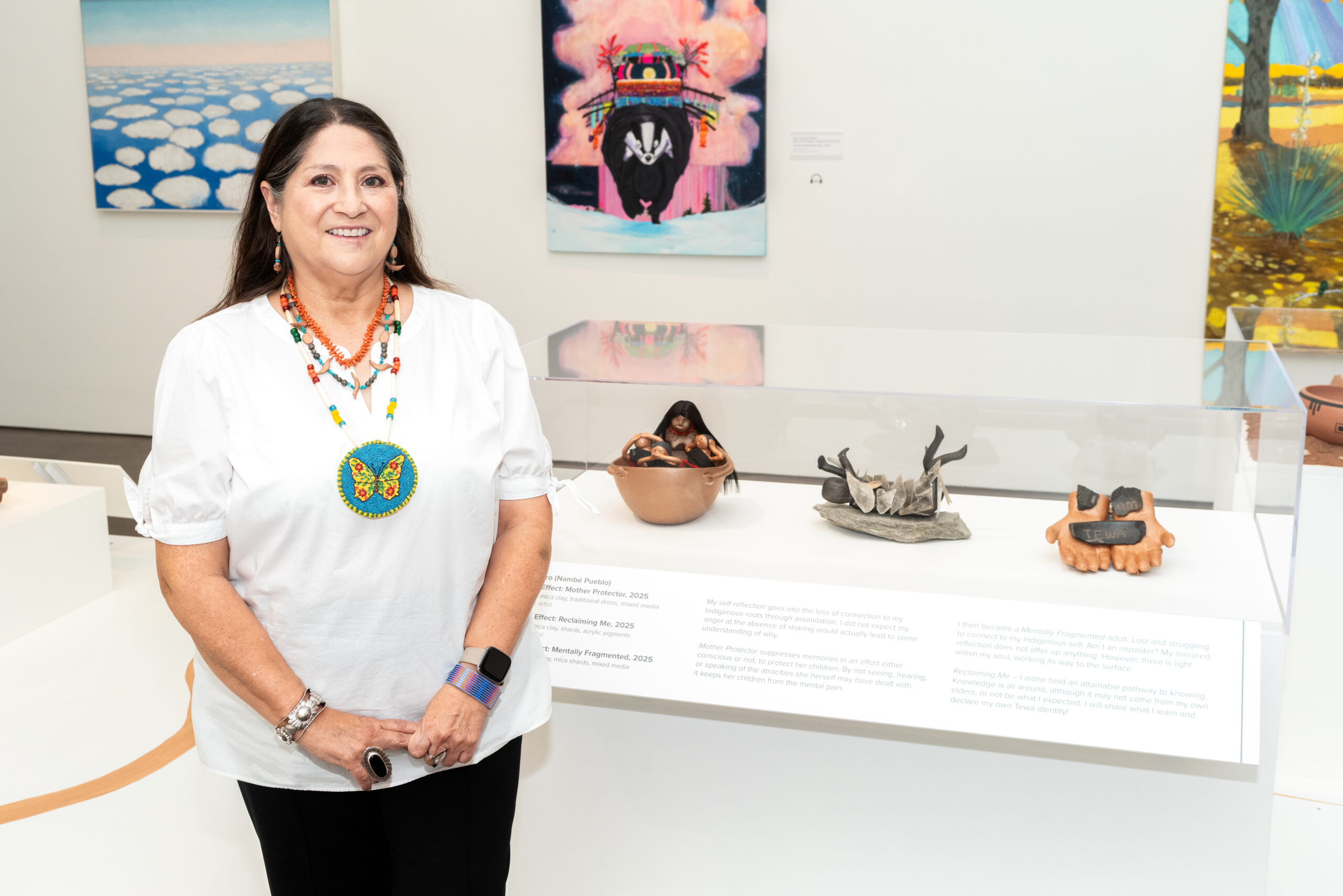 Photograph of a person in a white shirt and long dark hair standing and smiling in front of a case with several ceramic pieces displayed in a gallery.