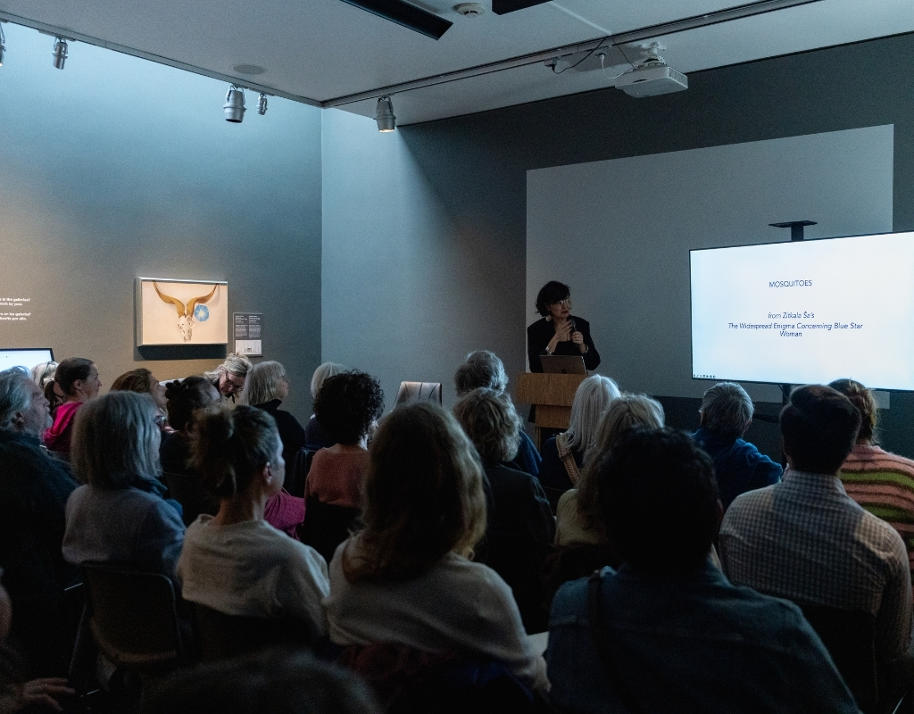 A person stands at a podium in a dark room with a screen to their left and a painting on the wall to their right. They speak to a large crowd of seated listeners.