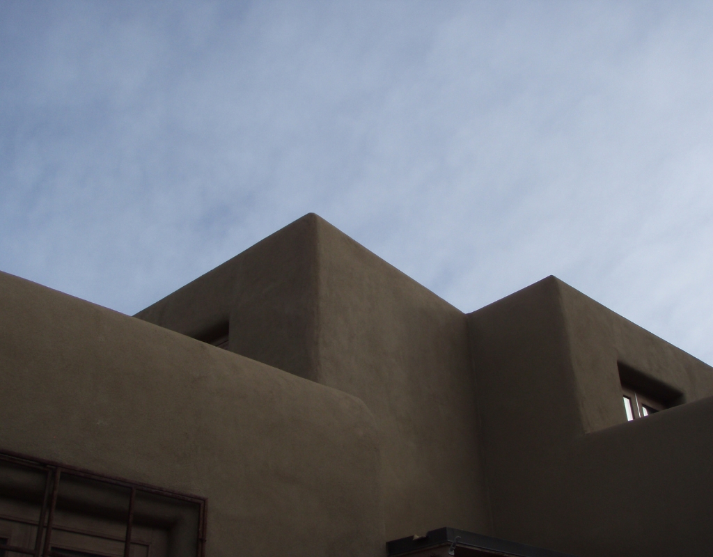 Photograph of the roof and upper walls of an adobe building with sharp pointed edges viewed from below. Behind the building is a blue sky with soft white clouds.