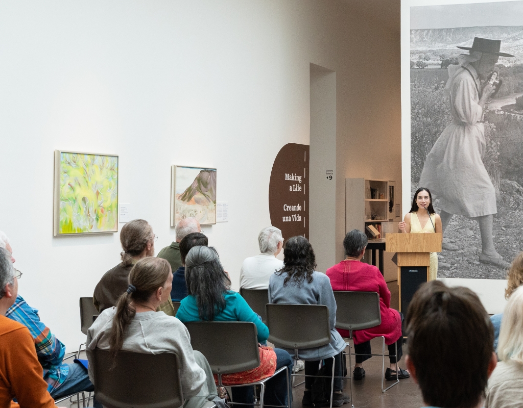 A person stands at a podium and address a crowd of people seated in a gallery with white walls.