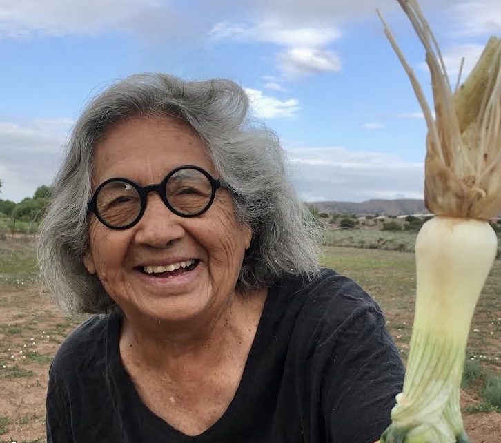 Photograph of a person with shoulder-length grey hair and glasses smiling at the camera while holding up a large vegetable. A blue sky is visible in the background. 