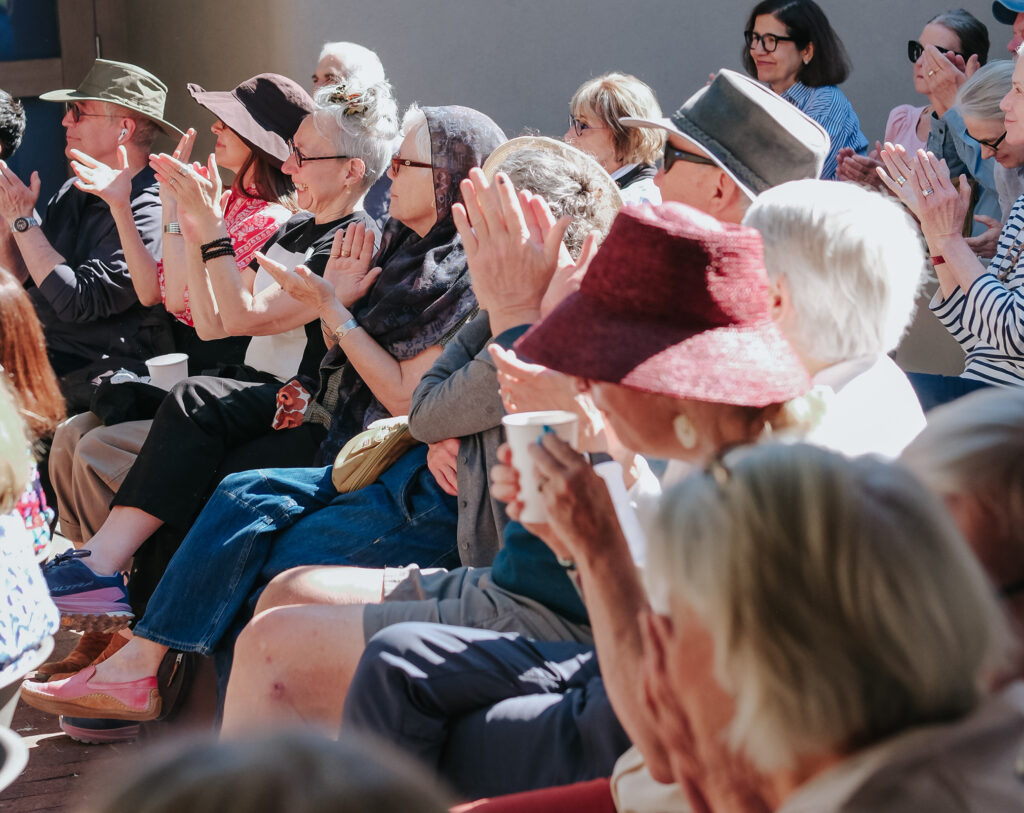 A group of people in an audience sit outdoors and clap. Several of them are wearing hats and sunglasses.