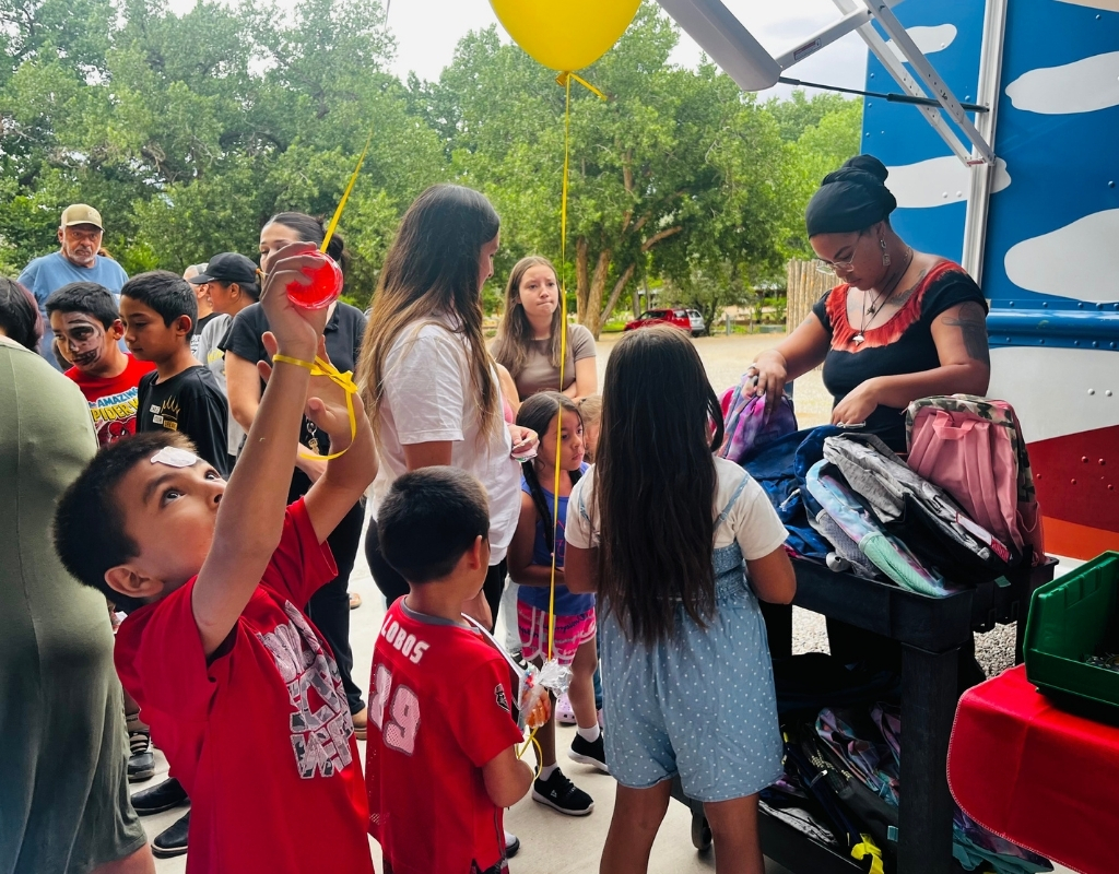 A crowd of young people, two holding balloons, line up as they receive backpacks.