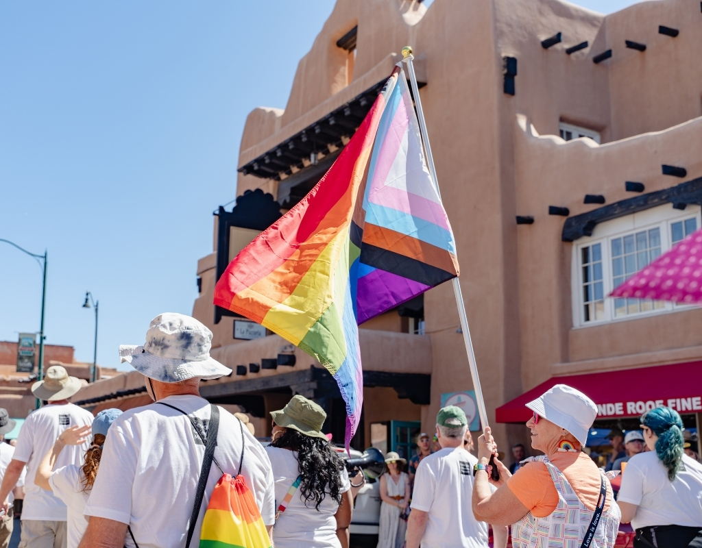 A group of people wearing white shirts with the Museum logo walk along a street in downtown Santa Fe. A person waves a large colorful Pride flag. Several people in the group have hats and colorful bags.