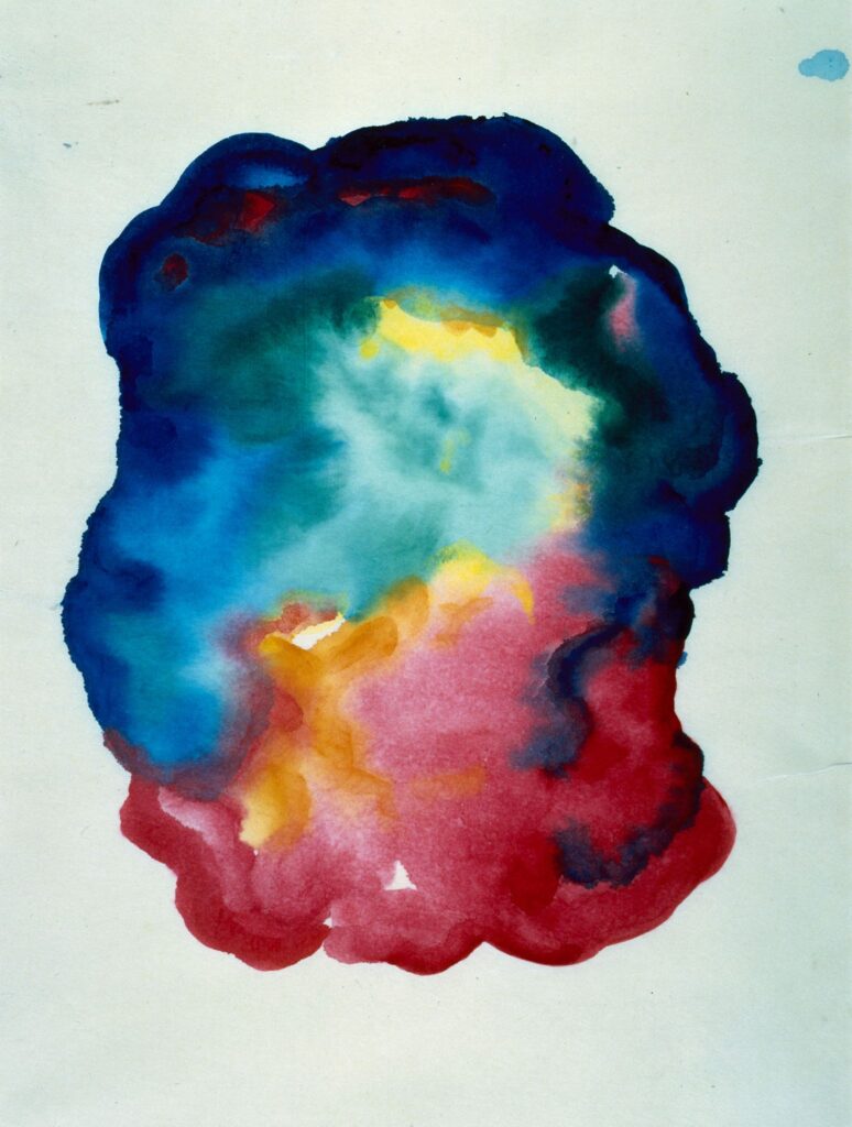 An amorphous blob of flowing blue, green, and red paint occupies the center of the paper. Patches of yellow dot the image. The form resembles a dark, colorful cloud, and contrasts with the plain white paper framing it.