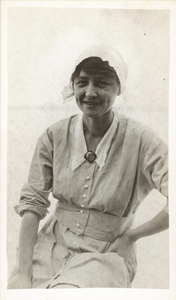 Black and white photograph of Georgia O'Keeffe wearing alight head covering and dress with a collar. O'Keeffe sits with one hand on her hip. Behind are pale plains.