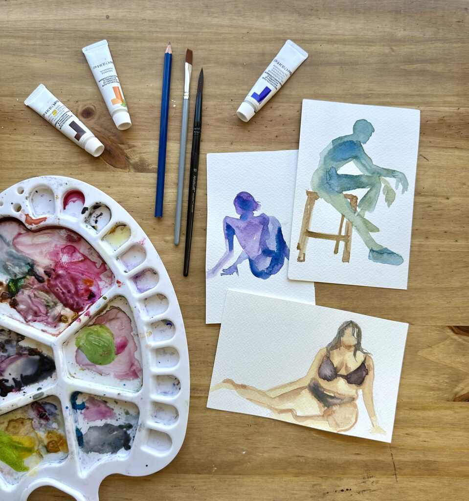 Three small watercolor paintings of figures in shades of blue, purple, and beige. Nearby on the same table rests a colorful watercolor palette and a few paint tubes and brushes.