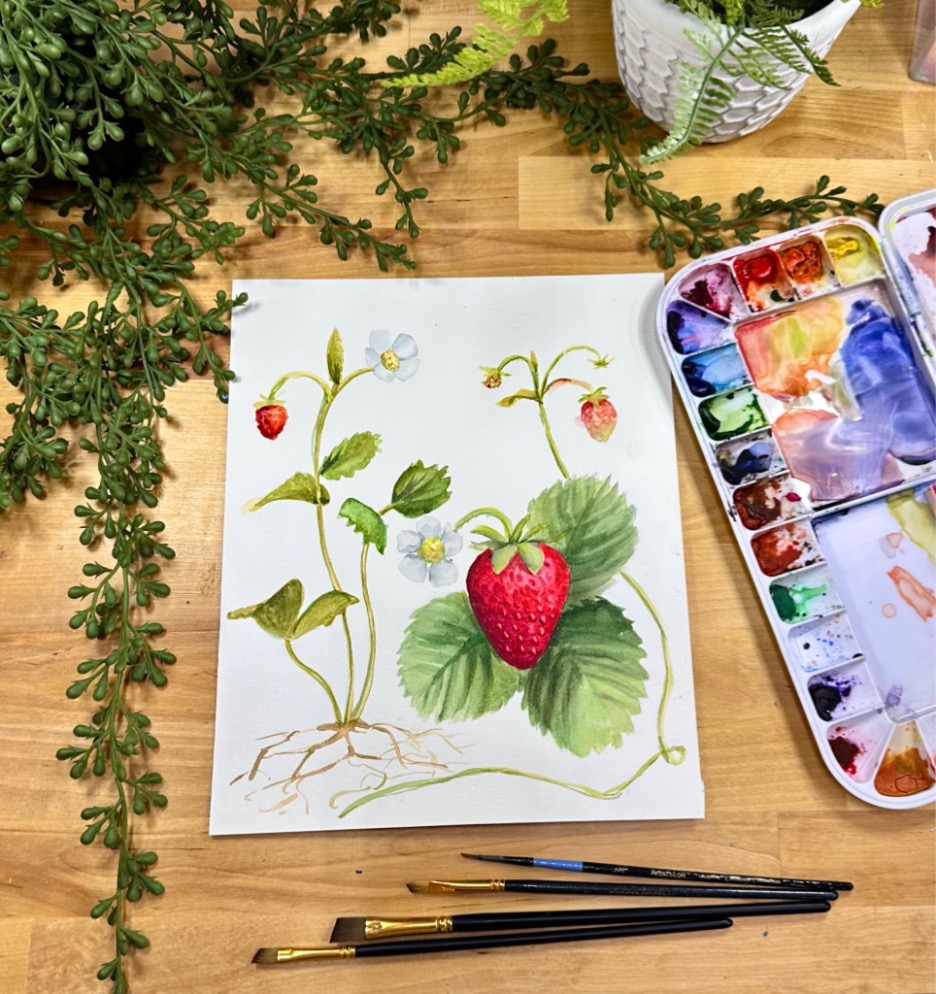 A painting of colorful strawberry plants. Nearby is palette of watercolors, small paintbrushes, and some green plants.