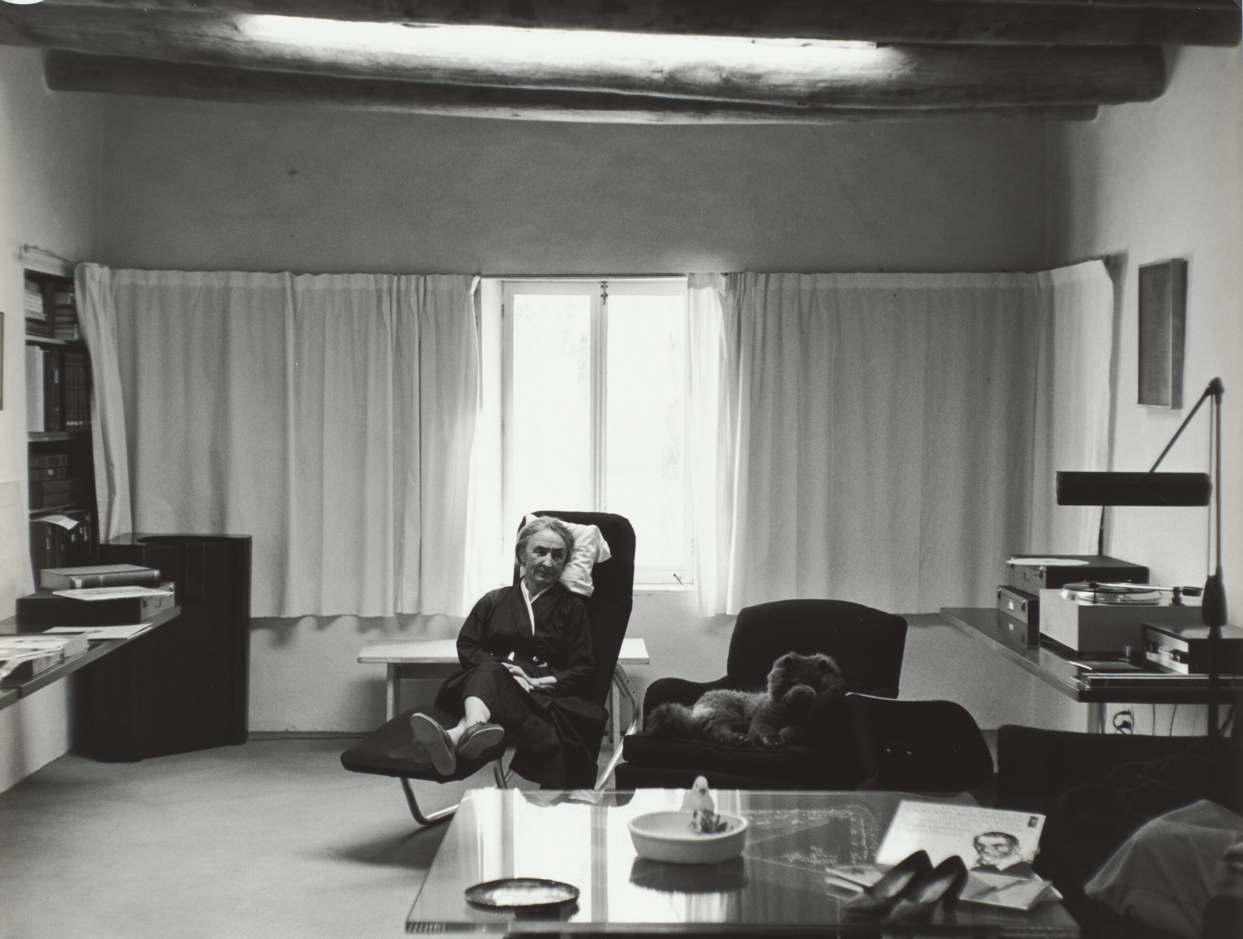 Black and white photograph of O'Keeffe seated in a lounge chair dressed in a black wrap dress with her Hector Aguilar belt, in the middle of a living room, with a wall of white drapes on the back wall, two of which are open right behind her. One of her Chow Chow dogs lies in the chair to her left. In front of them a glass coffee table with a pair of high-heels, plate, and record sleeve. To the right, a detail of a person's legs are visible on the sofa. In the back right, a table with stereo and a large lamp, in the back left a ledge and shelves with papers.