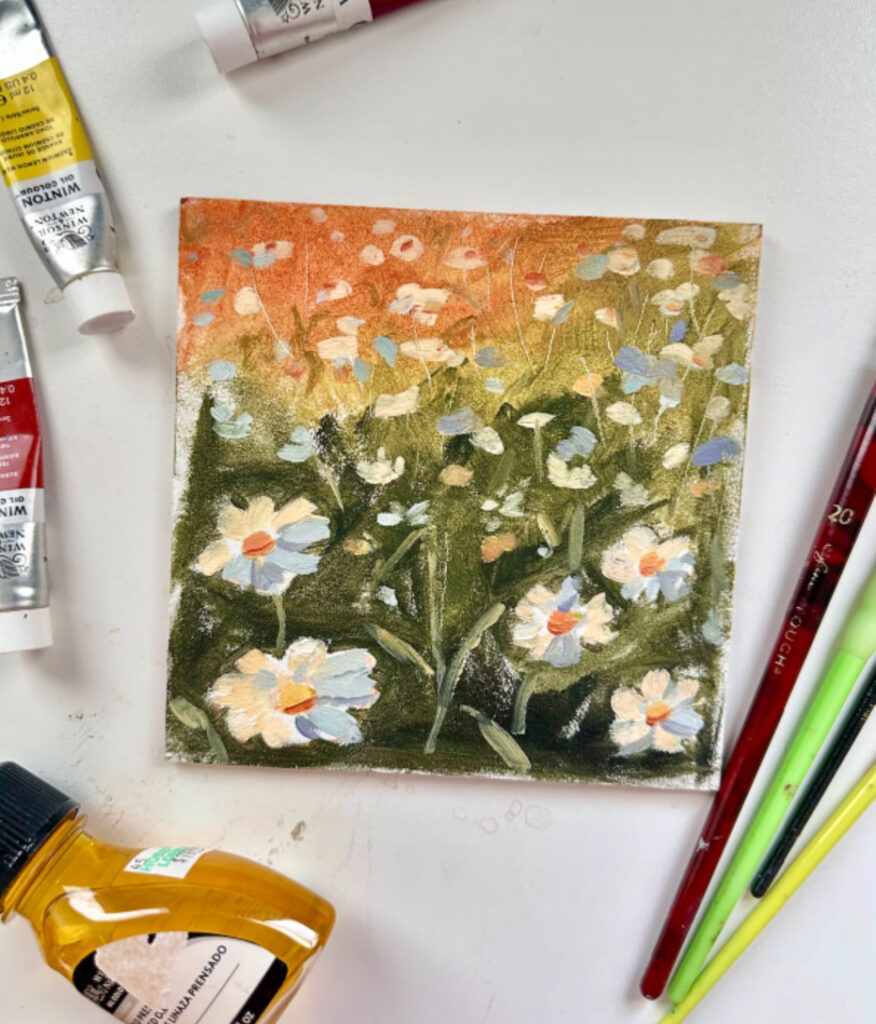 A small canvas with oil paintings of white daisies against a green and orange background. Surrounding the small canvas are paint tubes and several paintbrushes.