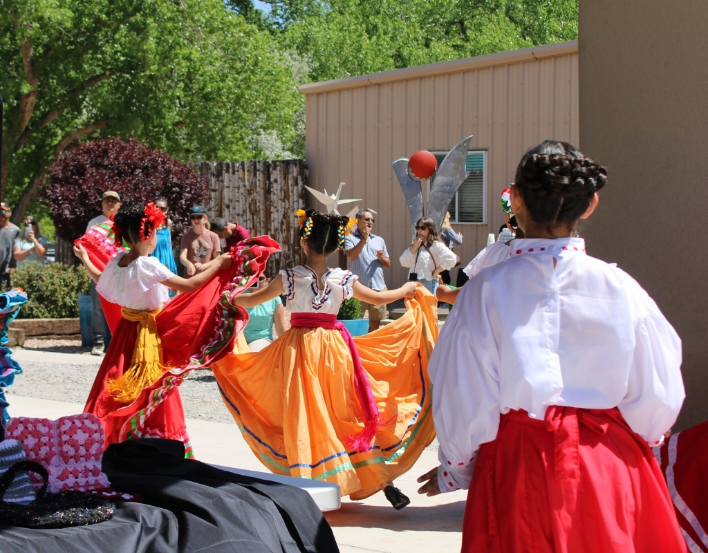 A group of young dancers with colorful large skirts perform in front of a crowd outside.