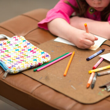 Close-up photograph of a child drawing on a piece of paper with colored pencils on top of a leather bench. The child’s face is out of frame and focus is on their hands, a patterned pencil and the colored pencils scattered over the bench. A second child’s hand pokes in from the right side of the frame.