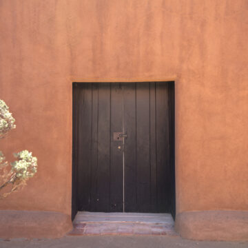 This photograph features the outside patio and door at Georgia O'Keeffe's Abiquiu Home and Studio. Foliage is visible on the left side of the frame.