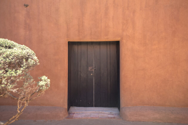 This photograph features the outside patio and door at Georgia O'Keeffe's Abiquiu Home and Studio. Foliage is visible on the left side of the frame.