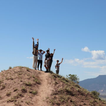 Photo of a group of young students on a hill with a blue sky and Pedernal mountain behind them