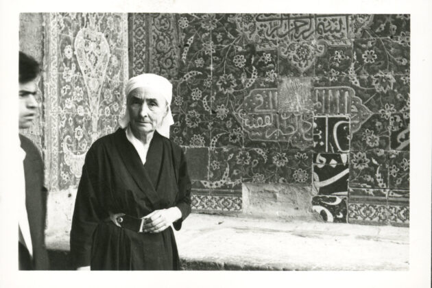 Black and white photograph of Georgia O’Keeffe wearing a dark wrap dress and a light scarf around her head at a Mosque. Behind her are the intricate tiles of the Mosque wall and to the left of the frame is gentleman passing through in front of the camera.
