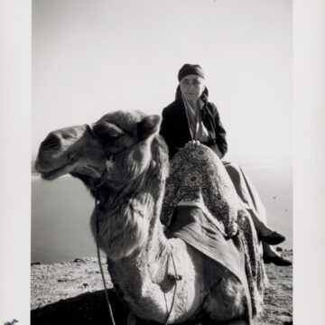 A black and white photograph of Georgia O'Keeffe seated on a camel. The camels face looks to the left in the foreground. Georgia O'Keeffe sits side saddle and is draped in fabrics with simple pumps on her feet.