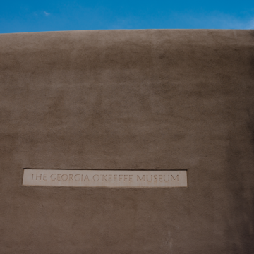 Adobe wall exterior of the Georgia O'Keeffe Museum with a sign of the Museum's name. Above the wall is a sliver of blue sky and a little cloud