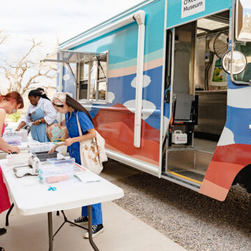 On the right is the Art to G.O. Truck with cloud motifs and an open window facing a few tables. On the left of the frame are tables with art supplies and a group of people standing around them making collages. In the background are trees.