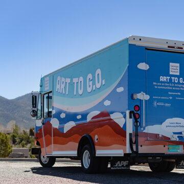 Image of the Museum's mobile creative studio with O'Keeffe-inspired clouds and mountains with the name Art to G.O. on it. The truck faces away from the camera and behind are mountains