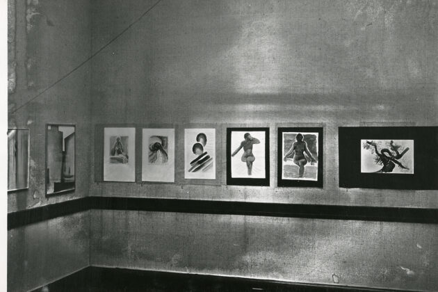 Black and white photograph showing several of O'Keeffe's watercolors on display. On the wall in focus are eight of O'Keeffe's watercolors, including a wall of an exhibition with three nude watercolors and several more. Under the hanging paintings is a dark moulding.