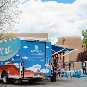Photograph of the 'Art to G.O.' truck with cloud motifs on the side in a parking lot under a tree. To the right of the truck and under it's awning, people are gathered over a table for art activities. Blue foam blocks with children playing in them take up the right side of the frame and behind the truck are adobe buildings and large white clouds.