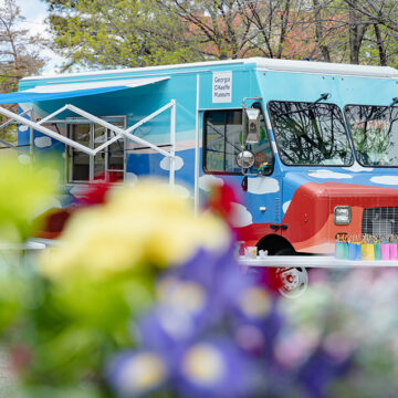 Photograph of the 'Art to G.O.' truck with cloud motifs on the side in a parking lot under trees. the bottom of the frame and front of the truck are partially covered with our of focus/ blurry flowers.