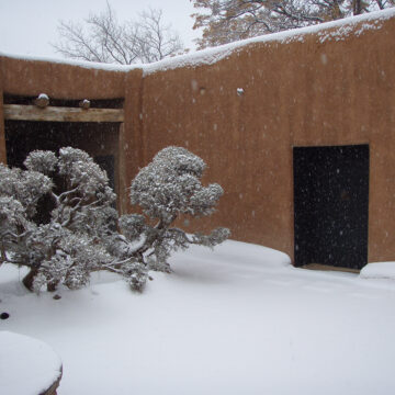 Photograph of an interior courtyard with a dark door on the right and square opening on the left with beams above. In the courtyard is the top of a circular well and a small trees. The entire space is covered in white snow.