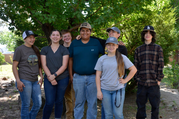 Group of 7 young people in long pants, some wearing baseball caps standing in a line smiling at the camera. Behind them are green trees.