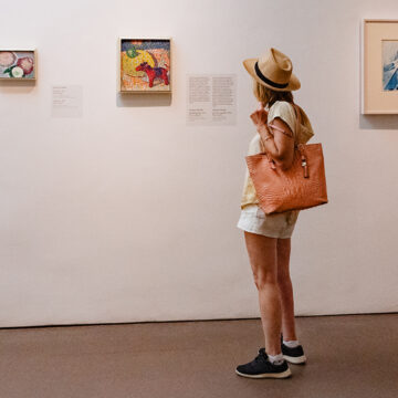 Photograph of a person in a hat and holding a bag looking at three small paintings on a white gallery wall.