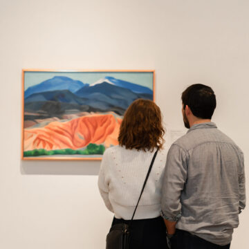 A photograph of two people taken from behind, they are standing close together looking at O’Keeffe’s painting ‘Black Mesa Landscape, New Mexico / Out Back of Marie’s II’ in the Museum galleries.
