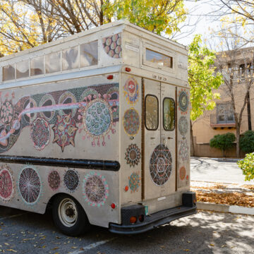 Photograph of a rectangular grey truck, a mobile art gallery, with artwork on the sides and front. The truck is parked underneath leaves with golden foliage.
