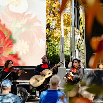 Three people in dark clothes holding and playing instruments including a guitar and a violin. Behind the is a large screen with a red paintings by O'Keeffe and surrounding the scene is fall folliage