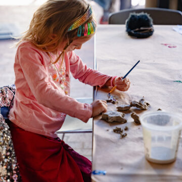 Photograph of a child sitting at a table making art with pieces of clay and a paintbrush.