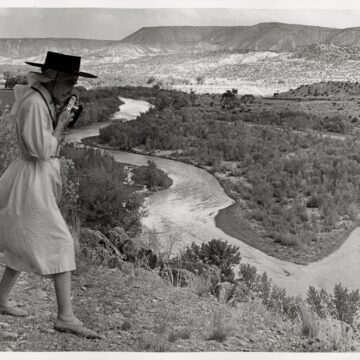 Portrait of O'Keeffe walking in the Chama Valley - a sprawling high desert landscape with the Chama River winding in front of mesa outcroppings against a big sky with clouds; full-length profile of O'Keeffe walking against the wind in the landscape while holding a 35 mm camera to her chin ( which could be a Leica) ; she wears a white head scarf under her black wide brimmed hat, a white wrap dress which, along with her head scarf, is blowing in the wind, and expensive light colored flat leather shoes